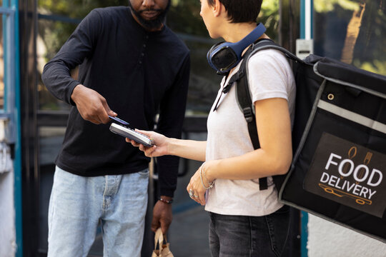 Female delivery employee courier has pos terminal for man to pay, client taps card on the wireless device for payment. Courier ensuring express service and easy transaction for takeaway.