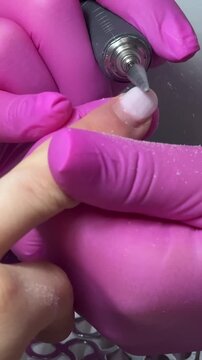 Young woman middle finger in focus as a nail technician wearing pink latex gloves removes old gel polish using an electric nail drill tool against a clean professional manicure setting.