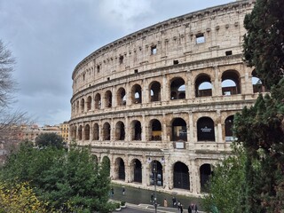 Fototapeta premium Rome, Italy - 11 January 2025. The Colosseum’s facade rises over trees and passersby, its arches weathered and grand beneath an overcast winter sky.