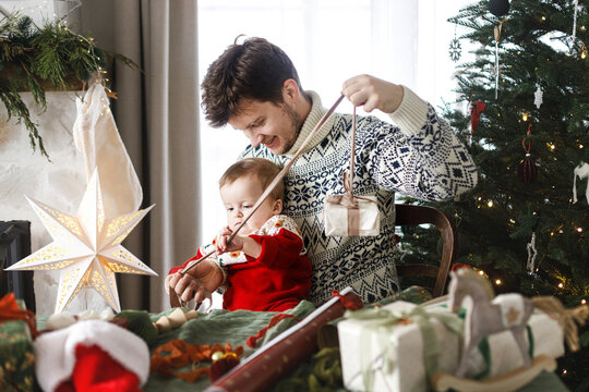 Merry Christmas! Happy father and baby son in stylish festive sweaters wrapping together christmas presents on background of modern decorated tree. Joyful family time, happy holidays - Powered by Adobe