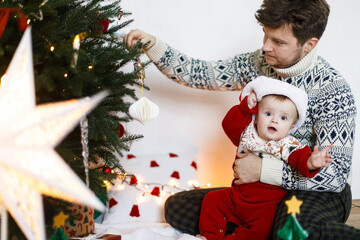 Merry Christmas! Happy father and cute baby son in santa hat and festive sweater decorating together christmas tree with modern bauble. Joyful family time, winter holidays preparation