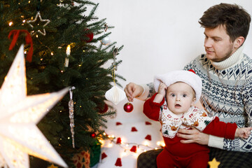 Merry Christmas! Happy father and funny baby son in santa hat and festive sweater decorating together christmas tree with modern bauble. Joyful family time, winter holidays preparation