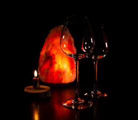 Two elegant empty clear wineglasses for red wine on the table with burning candle on dark black background with blur orange salt lamp. Closeup decor celebration