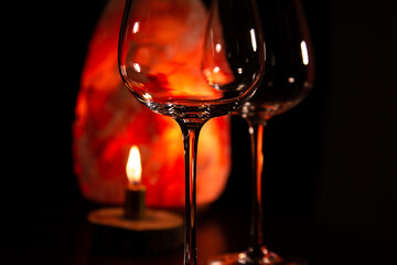 Closeup two elegant empty clear wineglasses for red wine on the table with burning candle on dark black background with blur orange salt lamp. Still life beautiful bright decor celebration
