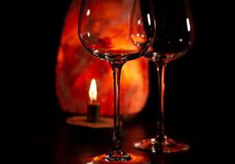 Two elegant empty clear wineglasses for red wine on the table with burning candle on dark black background with blur orange salt lamp. Closeup decor celebration