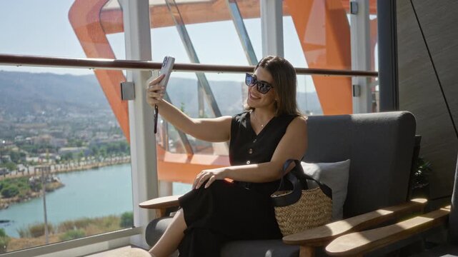 Woman holding smartphone on cruise terrace, smiling while seated by glass railing and taking a selfie; relaxation travel leisure.