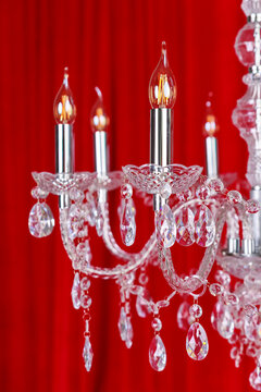A close-up of an elegant crystal chandelier with glowing bulb-shaped lights, showcasing intricate glass droplets and ornate arms against a rich red curtain backdrop
