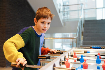 Young boy in colorful sweater playing foosball table game inside modern building. An enthusiastic...