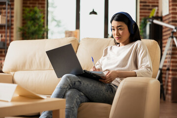Determined female freelancer sitting on couch with headphones, reviewing project presentation...