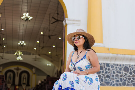 A woman in a blue and white dress sits on the steps of a church, wearing a straw hat and sunglasses.