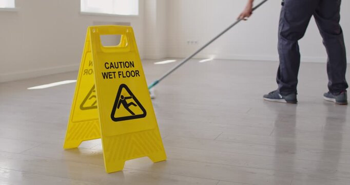 Janitor mopping floor in empty room with yellow caution wet floor sign placed in foreground. Worker cleaning slippery surface and ensuring safety by warning about freshly cleaned and wet area.
