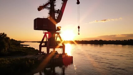 A large crane stands prominently at the riverbank, silhouetted against the bright sunset. The crane is positioned to lift materials as the sun sets over the calm river and landscap