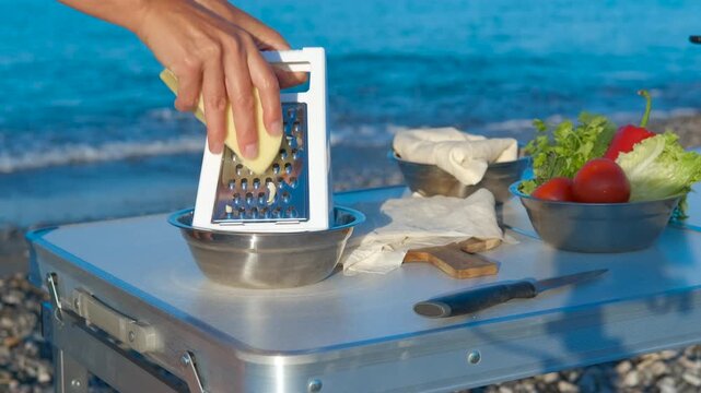 Woman grating cheese on beach for delicious meal preparation. Female grating cheese into a bowl on a portable table on a beach, with fresh vegetables and pita bread nearby, preparing a meal by the sea