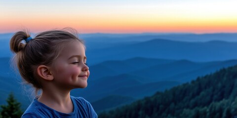 A young girl is smiling and looking out over a mountain range. The sky is a beautiful mix of orange and blue hues, creating a serene and peaceful atmosphere