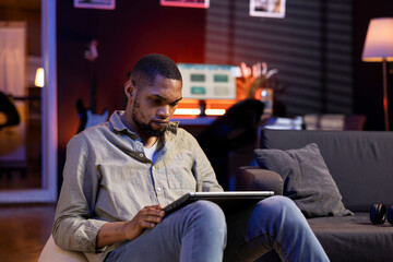 Focused african american guy multitasking on laptop in stylish living space, connected to the internet while managing his freelance work and remote responsibilities. Browsing web laying on sofa.