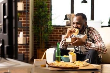 Portrait of man holding delicious burger, eating takeaway food and watching entertainment movie on television. African american male sitting on couch, enjoying tasty takeout during weekend at home.