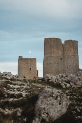Close-up view of the medieval fortress of Rocca Calascio in Abruzzo, perched on a rocky mountain ridge. Ancient stone walls and towers stand against a dramatic cloudy sky with the mountains.