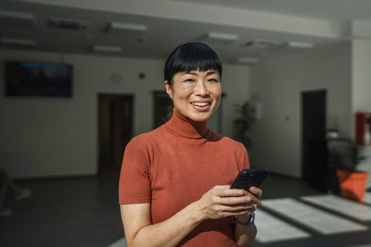 Asian woman smiling using smart phone in modern workspace