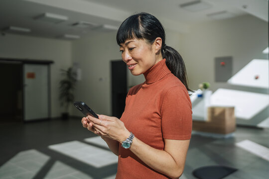 Asian woman smiling using smart phone in modern workspace - Powered by Adobe