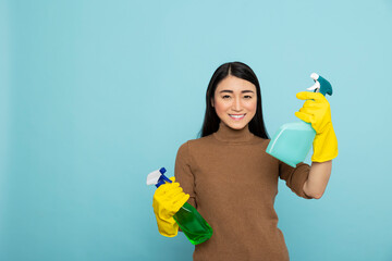 Excited and smiling asian janitor holding cleaning sprays in yellow rubber gloves, ready to start daily chores. Optimistic housewife with disinfectants, showing positive attitude toward housekeeping.