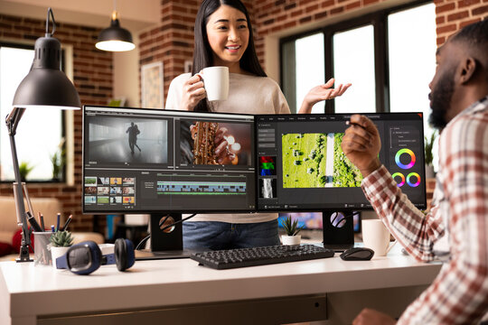 Diverse creative couple discussing footage and enjoying teamwork in cozy home studio. Asian woman and african american man collaborating on post production movie project with creativity and focus.