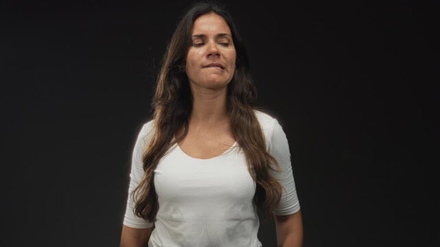 Woman biting lower lip showing bare neckline and chest while wearing a white shirt in studio with a black backdrop; skepticism.