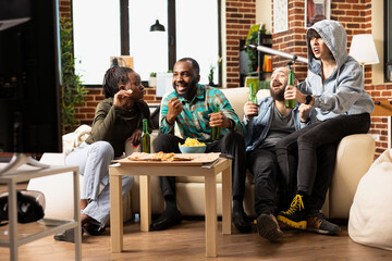 Joyful multiethnic friends bonding over TV entertainment in modern apartment. African American and Caucasian couples watching television together, sharing chips and beer in relaxed living room.