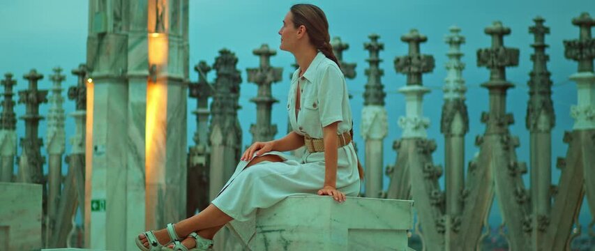 Woman sitting on marble ledge surrounded by ornate rooftop cathedral spires in Milan, italy