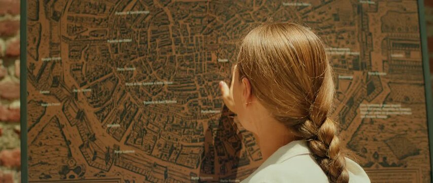 Back view of woman pointing at large vintage city map mounted on wall outdoors