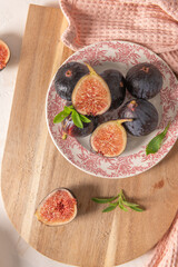 A close-up, top-down view of whole and halved figs on a decorative plate and a wooden board with stevia leaves and a pink napkin