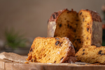 A warm close-up of a generous slice of traditional Italian Panettone with raisins and powdered sugar, set on a wooden board with the whole cake blurred in the background
