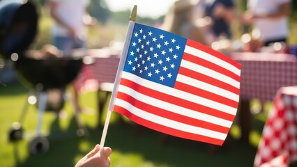 A hand holding a small American flag at a sunny outdoor barbecue.