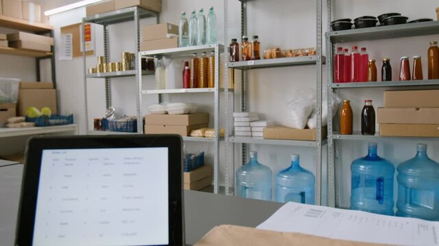 Medium shot of digital tablet with customer order on screen, brown paper packaging bags with delivery stickers. Groceries, bottles of drinks, canned foods stacked on racks in background