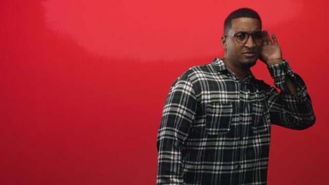Young black man in plaid shirt cupping ear with hand listening in studio against red backdrop; curiosity.