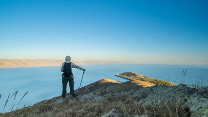 A hiker with a backpack standing on a mountain ridge overlooking Lake Sevan, Armenia. Stunning panoramic view of blue water, hills, and clear sky symbolizing adventure and freedom.