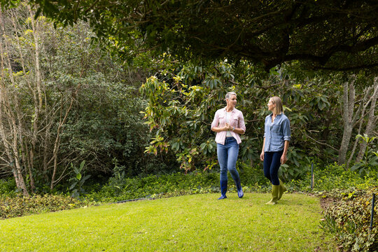 Female friends walking across lawn in home garden wearing casual shirts and rubber boots