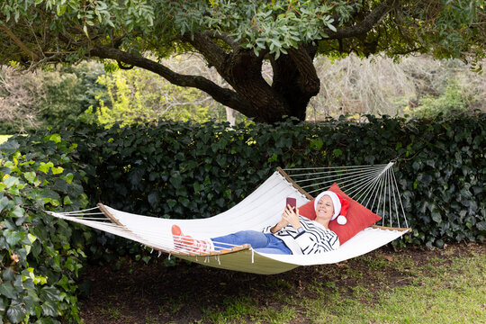 Woman reclining on hammock under tree in garden wearing Santa hat and red socks holding smartphone