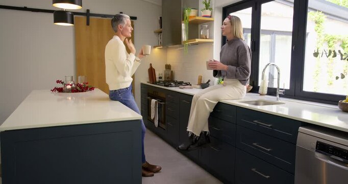 Female friends greeting, exchanging coffee chat for casual catch up with mugs on kitchen island - Powered by Adobe