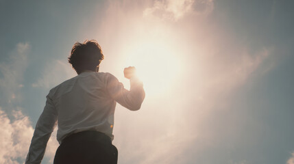 Young man back view wearing white shirt and black trouser raising fist toward bright sun with dramatic sky and lens flare expressing determination and hope