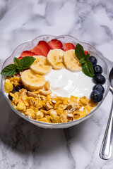 bol de vidrio con yogurt griego decorado con granola bananas frutillas arándanos y hojas de menta en fondo de mármol blanco