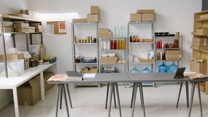 Full no people shot of interior of dark store fulfilling online orders, with various groceries on racks, packages and boxes on shelved, two desks with tablets for packing