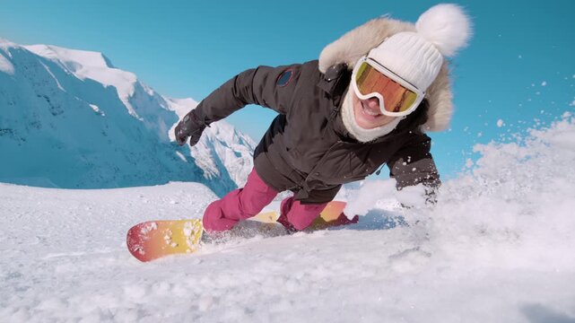 SLOW MOTION, CLOSE UP: Smiling snowboarder carves through snowy powder and crashes into soft snow. Young woman is learning to snowboard in high alpine mountains. Fun, authentic winter holiday vibe.