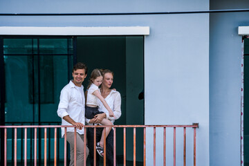 Obraz premium A happy family of three shares a loving moment on the balcony of their modern home. The daughter kisses her mother's cheek, showcasing joy, affection, and peaceful family life.
