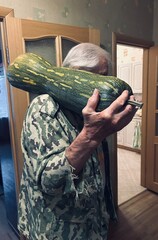 Senior man in camouflage jacket holds a large zucchini over his shoulder inside a cozy home. The image captures the relaxed and informal moment as he proudly displays the vegetable.