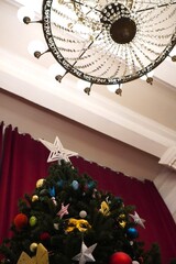 Christmas tree adorned with colorful ornaments and topped with a white star, viewed from below alongside a vintage chandelier, set against rich red curtains. Festive holiday atmosphere