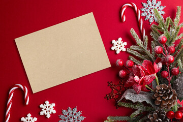 Christmas red background with blank paper, poinsettia, decorative fir branch, candy canes and cones for tree decoraton. New Year winter holiday composition with decor and space for text, top view.