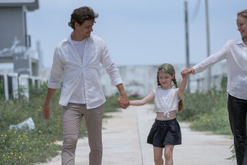 Fototapeta premium A cheerful family walks together through their new neighborhood. The smiling parents hold their daughter's hands, lifting her playfully as they enjoy a happy walk in front of their home.