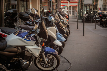 Row of parked motorcycles, motorbikes, and scooters outside a shop in France. Windshields and handlebars line up along the sidewalk, showing urban two wheeler mobility and retail context.