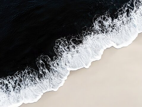 An aerial view captures the dramatic contrast between the dark, deep ocean water and the bright white foam of waves gently rolling onto a light-colored sandy be