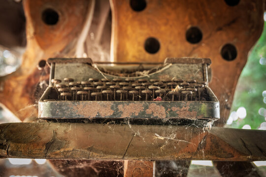 Close up of a rusty old typewriter covered in cobwebs and dust, sitting on a wooden surface, illustrating technological obsolescence and the passage of time in writing tools and office equipment.